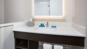Un baño con lavamanos blanco, tocador oscuro y espejo sobre el lavamanos.