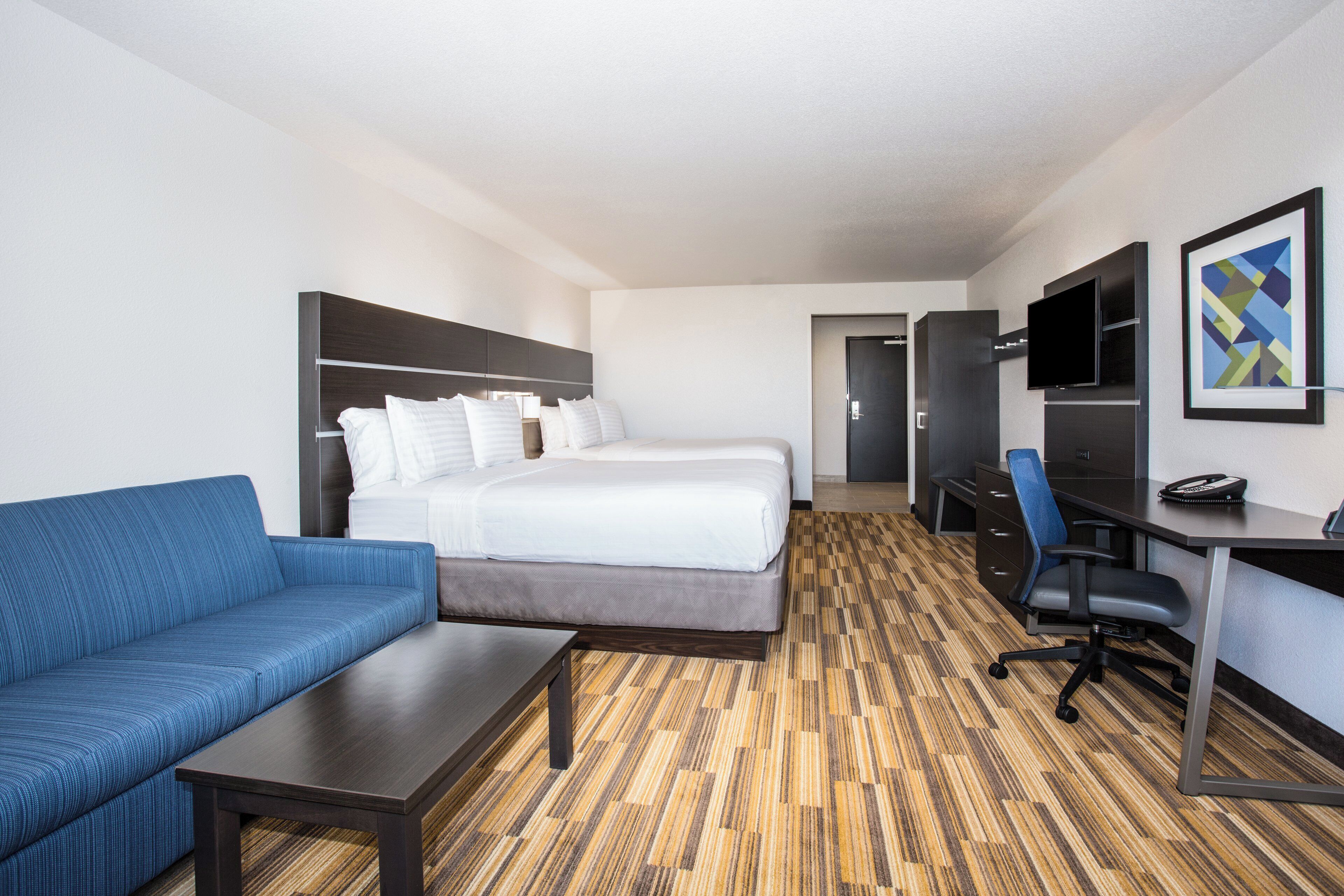 A hotel room with a bed, a sofa, a desk, a TV, and a painting on the wall.