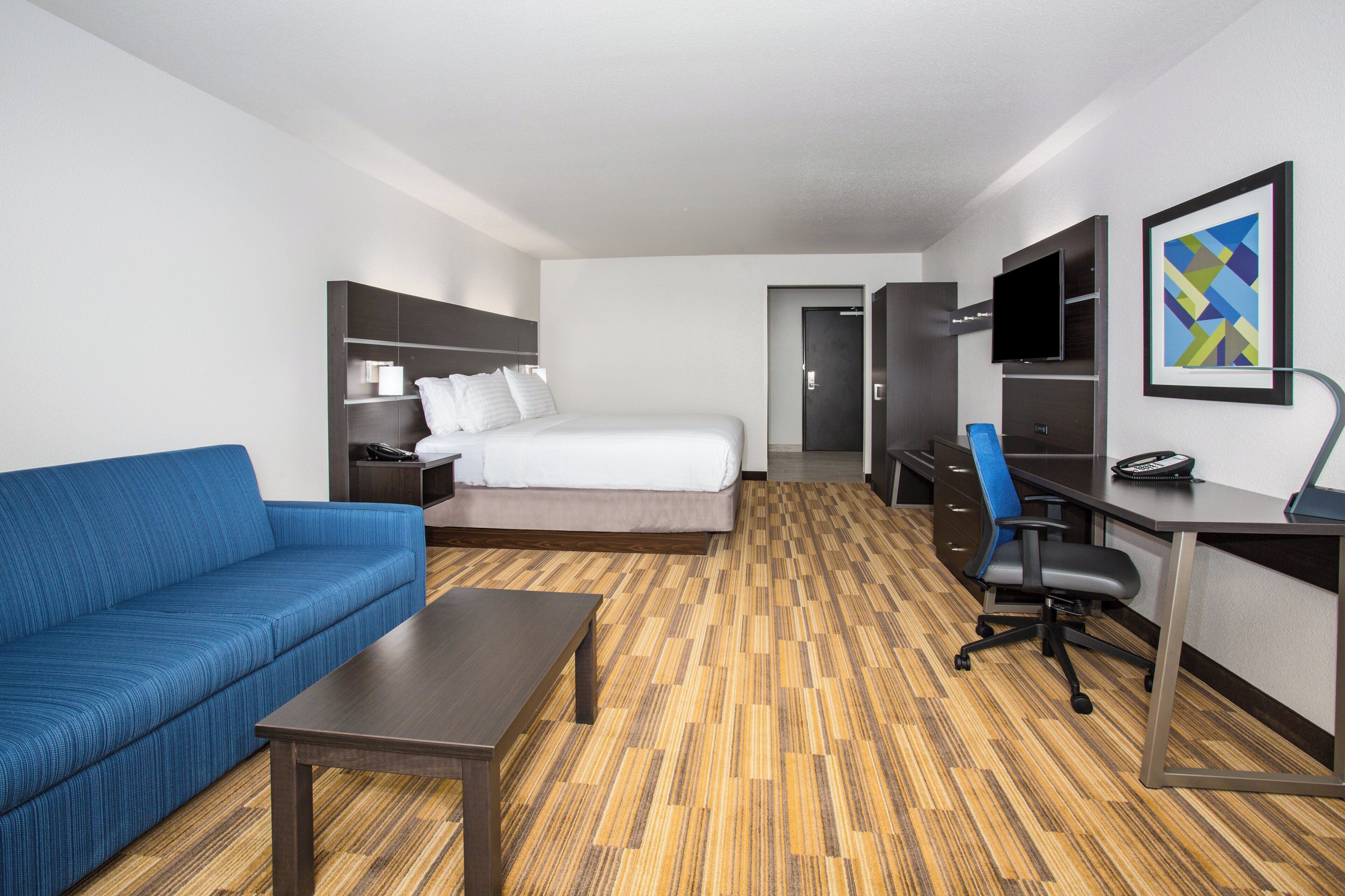 A hotel room with a bed, a blue sofa, a desk with a chair, a television, and a framed picture on the wall.