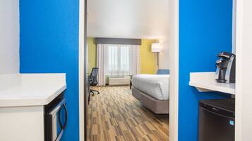 A hotel room with a bed, a desk, a chair, a coffee machine, and a refrigerator.