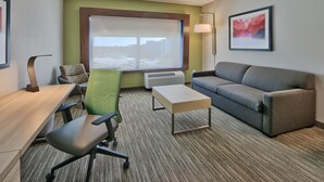 A hotel room with a desk, office chair, grey sofa, and a large window with a view of buildings.