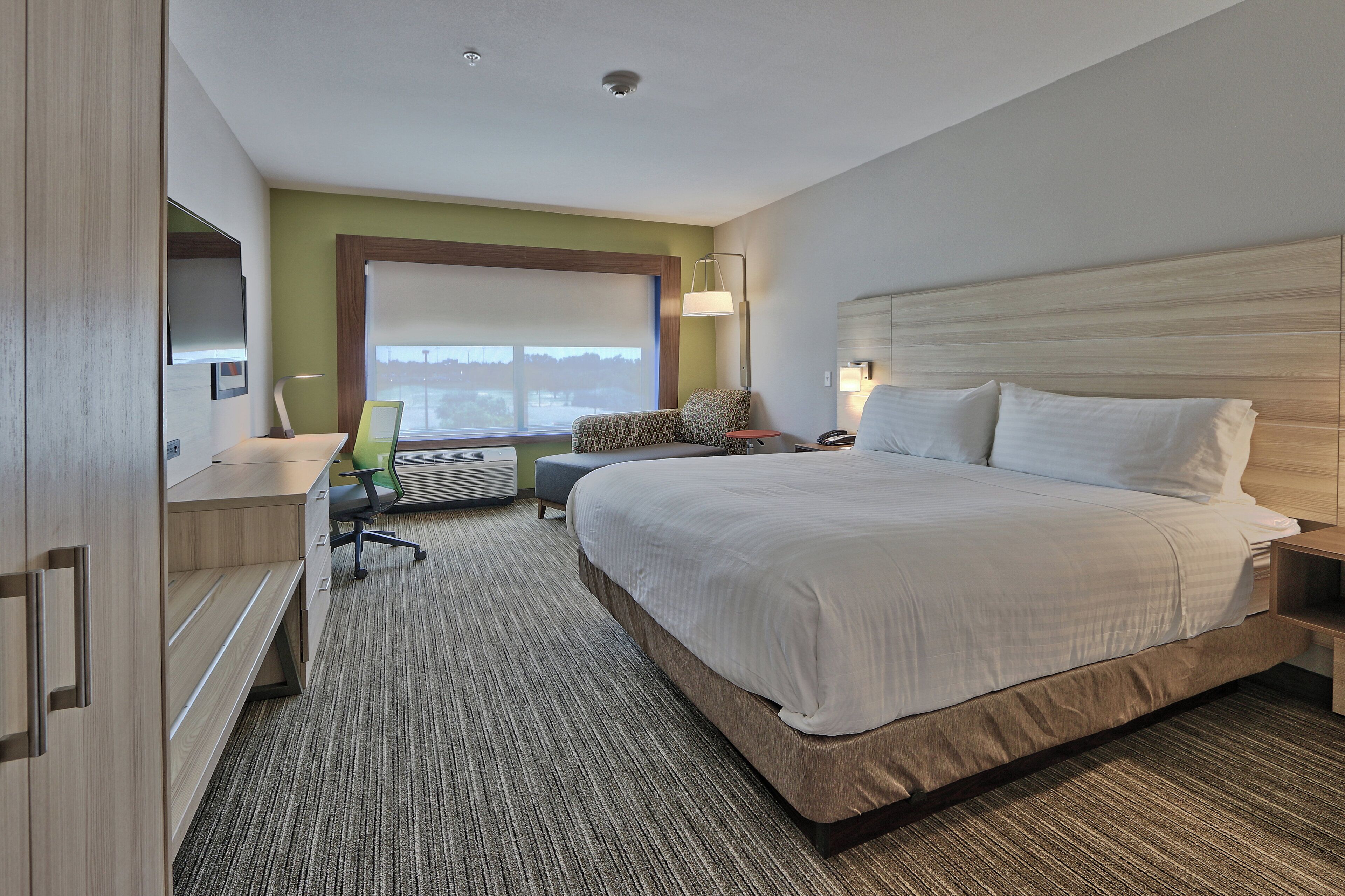 Une chambre d’hôtel avec un grand lit, un bureau avec une chaise, une fenêtre avec des stores et une lampe fixée au mur.