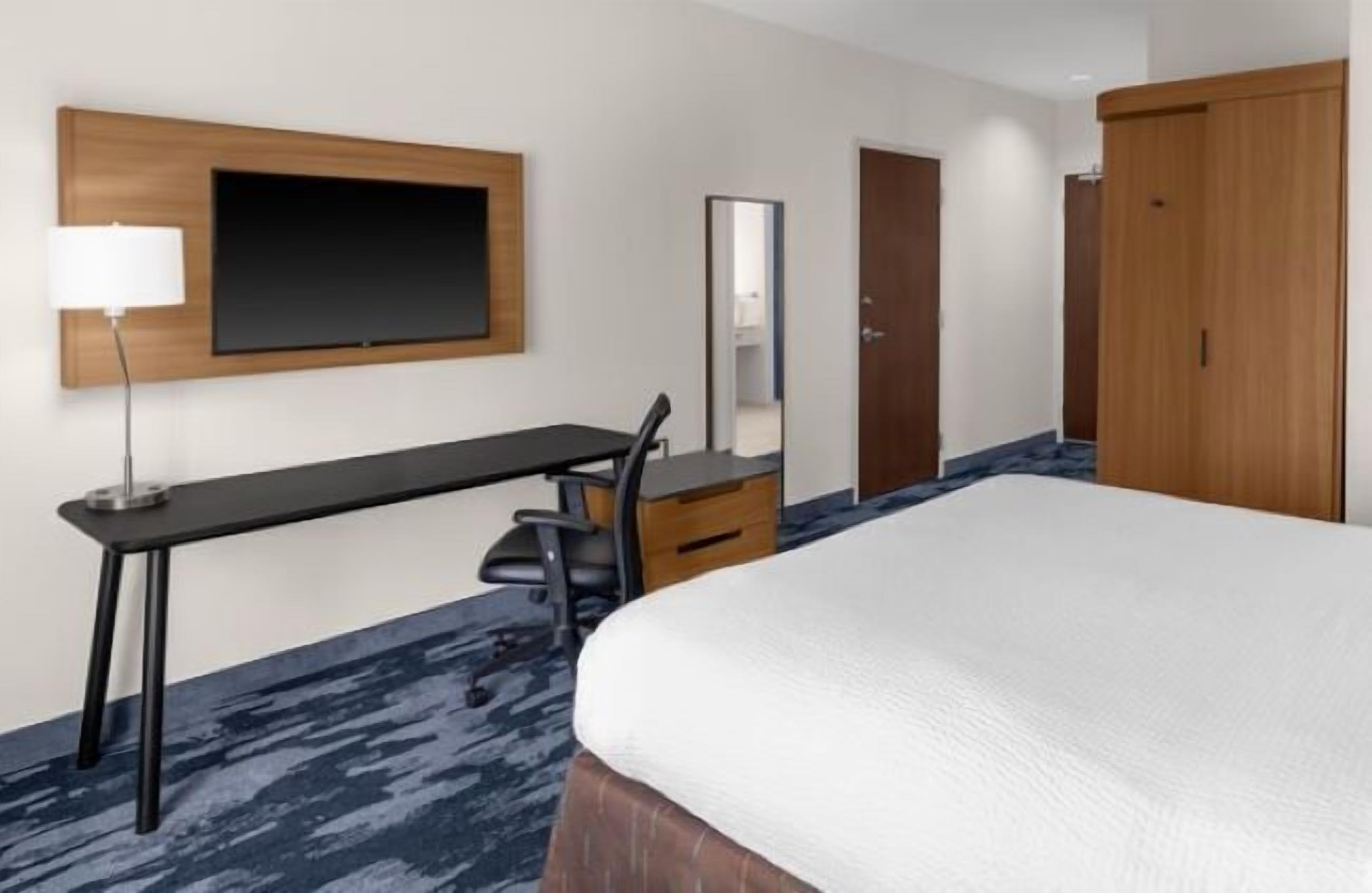 Habitación de hotel con cama, escritorio con silla, televisor montado en la pared, espejo y armario.