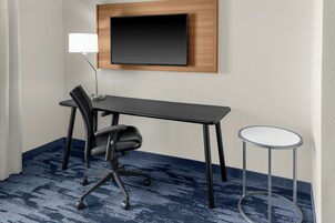 Habitación de hotel con un escritorio de madera oscura, una silla de oficina negra, una mesita auxiliar redonda blanca y un televisor de pantalla plana montado en la pared.