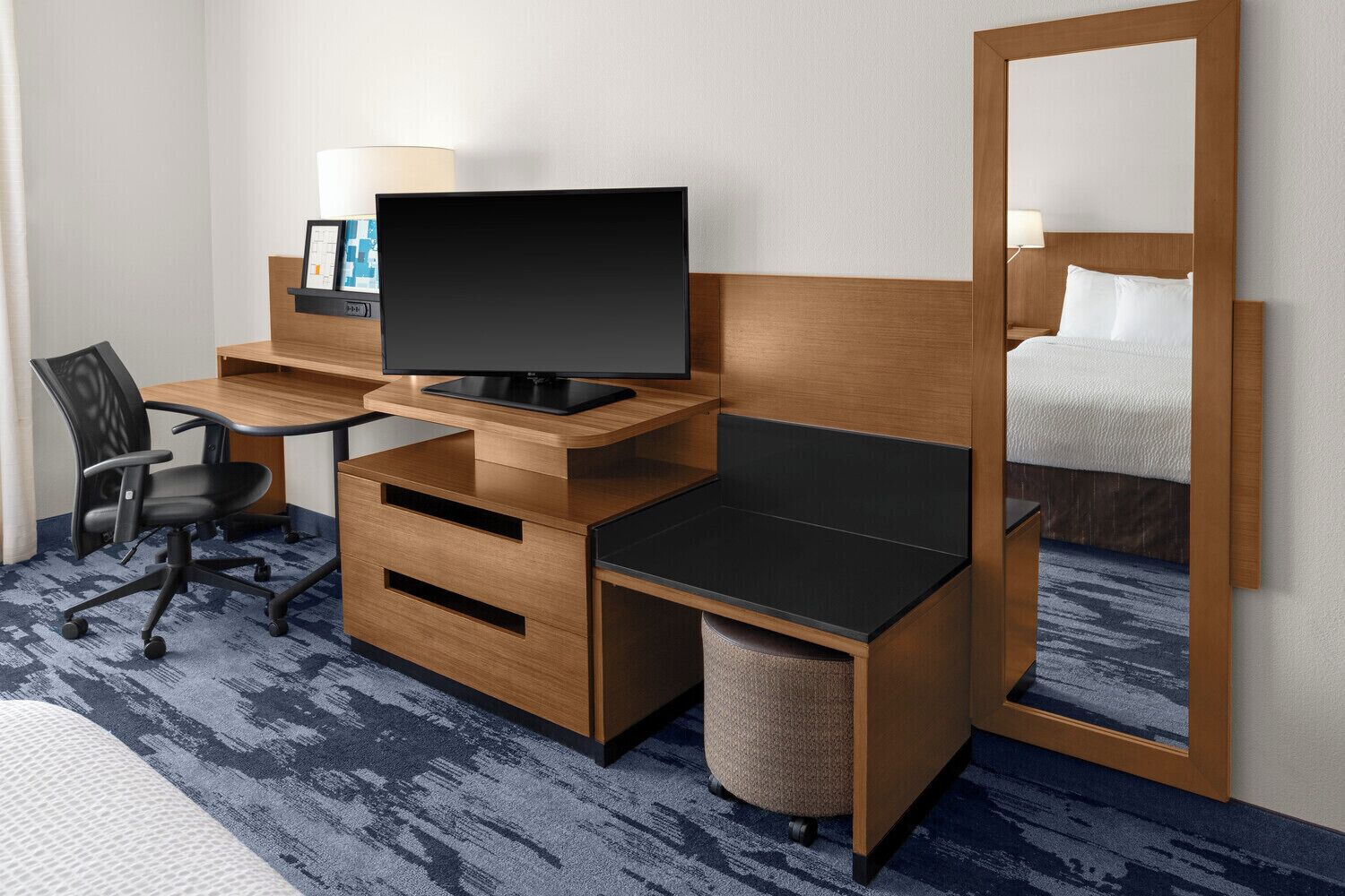 A hotel room with a wooden desk, a black chair, a television, a bed with white bedding, and a large mirror.