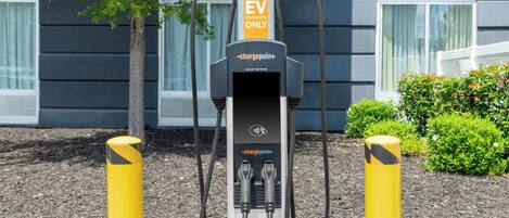 Een laadstation voor elektrische voertuigen met een bordje dat aangeeft dat het uitsluitend voor elektrische auto's is, gelegen voor een gebouw met ramen en een boom.