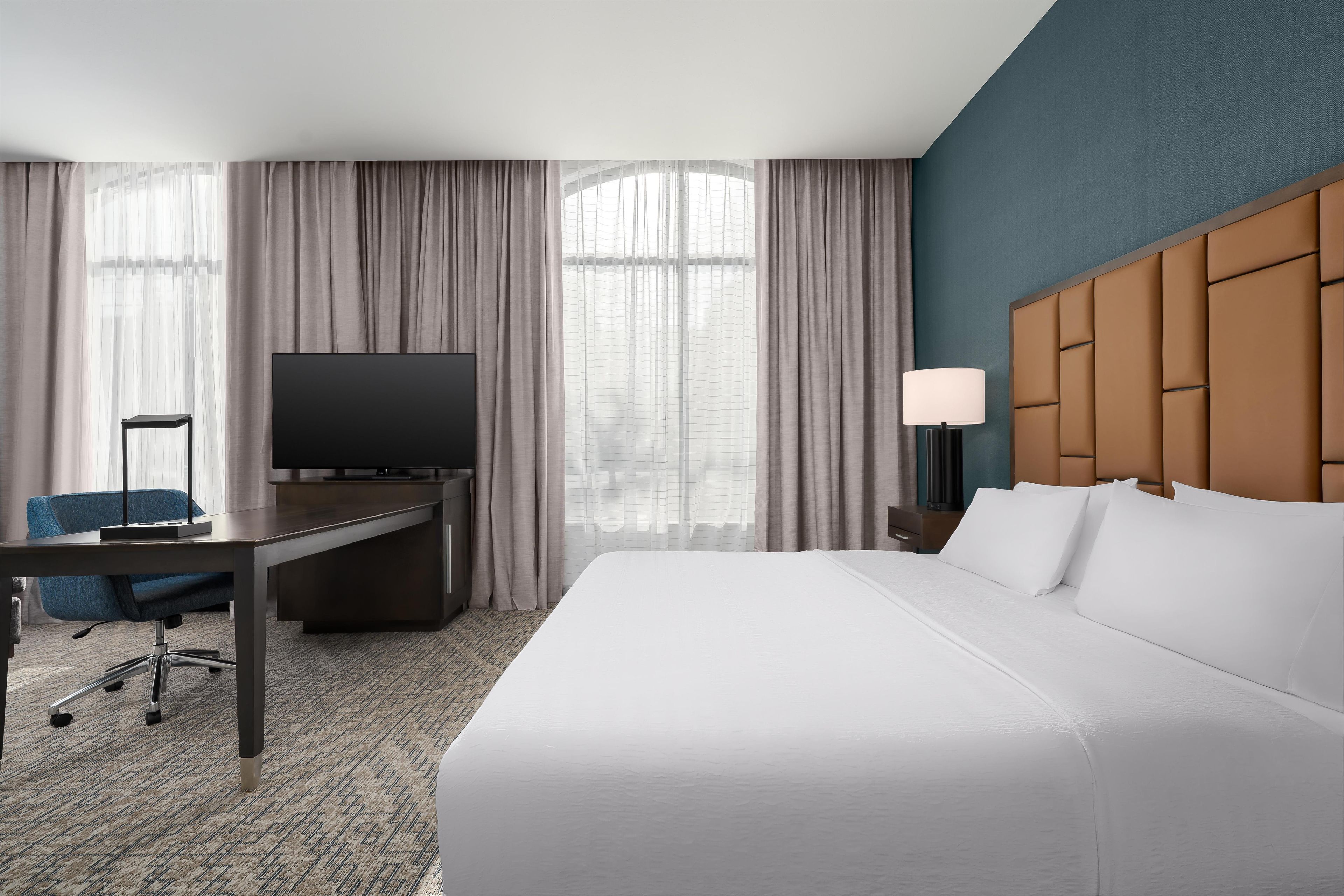 A hotel room with a large bed, a desk with a television, a chair, and a nightstand with a lamp.