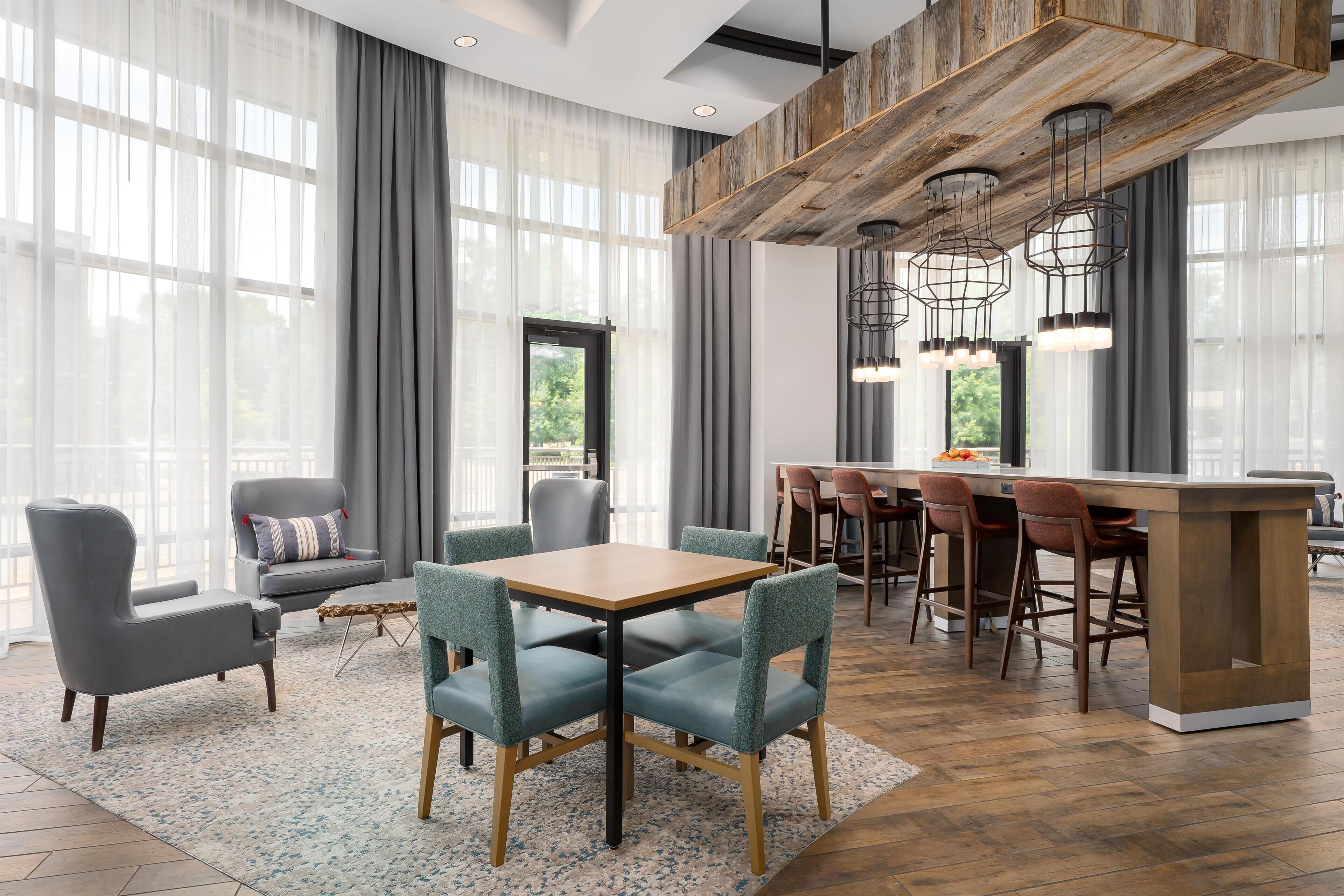 A modern hotel room with a dining area, large windows with sheer curtains, and a bar with stools.