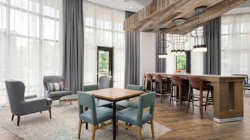 A modern hotel room with a dining area, large windows with sheer curtains, and a bar with stools.