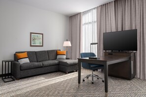 A hotel room with a gray sectional sofa, a desk with a flat-screen TV, a lamp, and a framed picture on the wall.