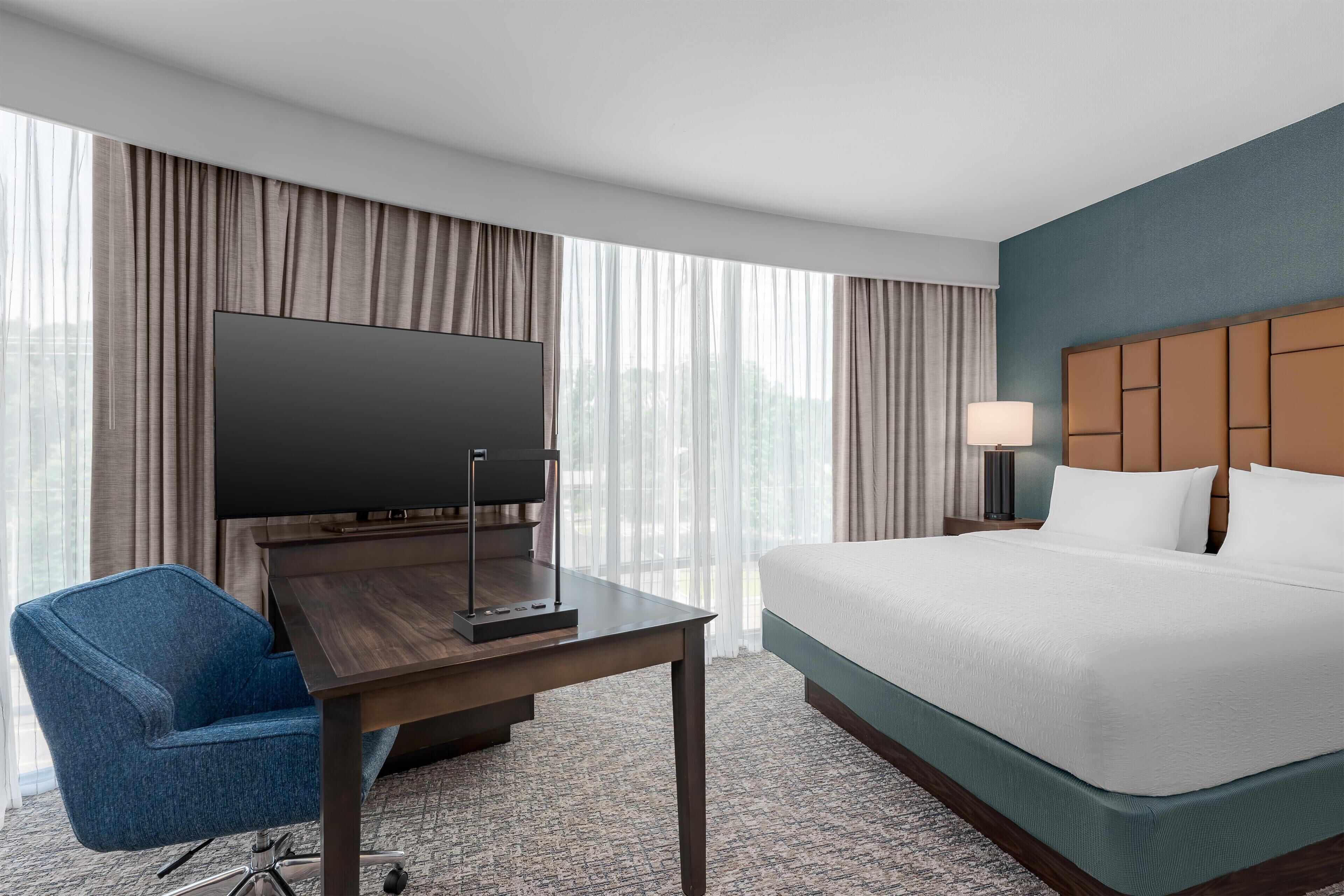 Une chambre d’hôtel comprenant un lit, un bureau avec un téléviseur à écran plat, une chaise, une lampe et une vue sur l’extérieur grâce à de grandes fenêtres.