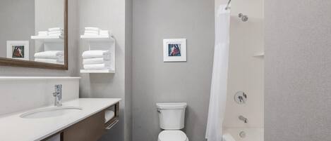 A modern bathroom with a white sink, toilet, and bathtub. There are towels stacked on a shelf, and a framed picture on the wall.