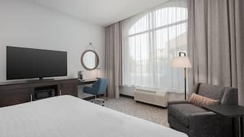 Habitación de hotel con una cama grande, un escritorio con silla, televisión, espejo, ventana con cortinas y lámpara.