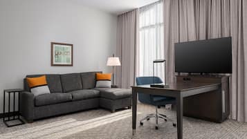 Habitación de hotel con un sofá seccional gris, un escritorio con televisor de pantalla plana, una lámpara y un cuadro en la pared.