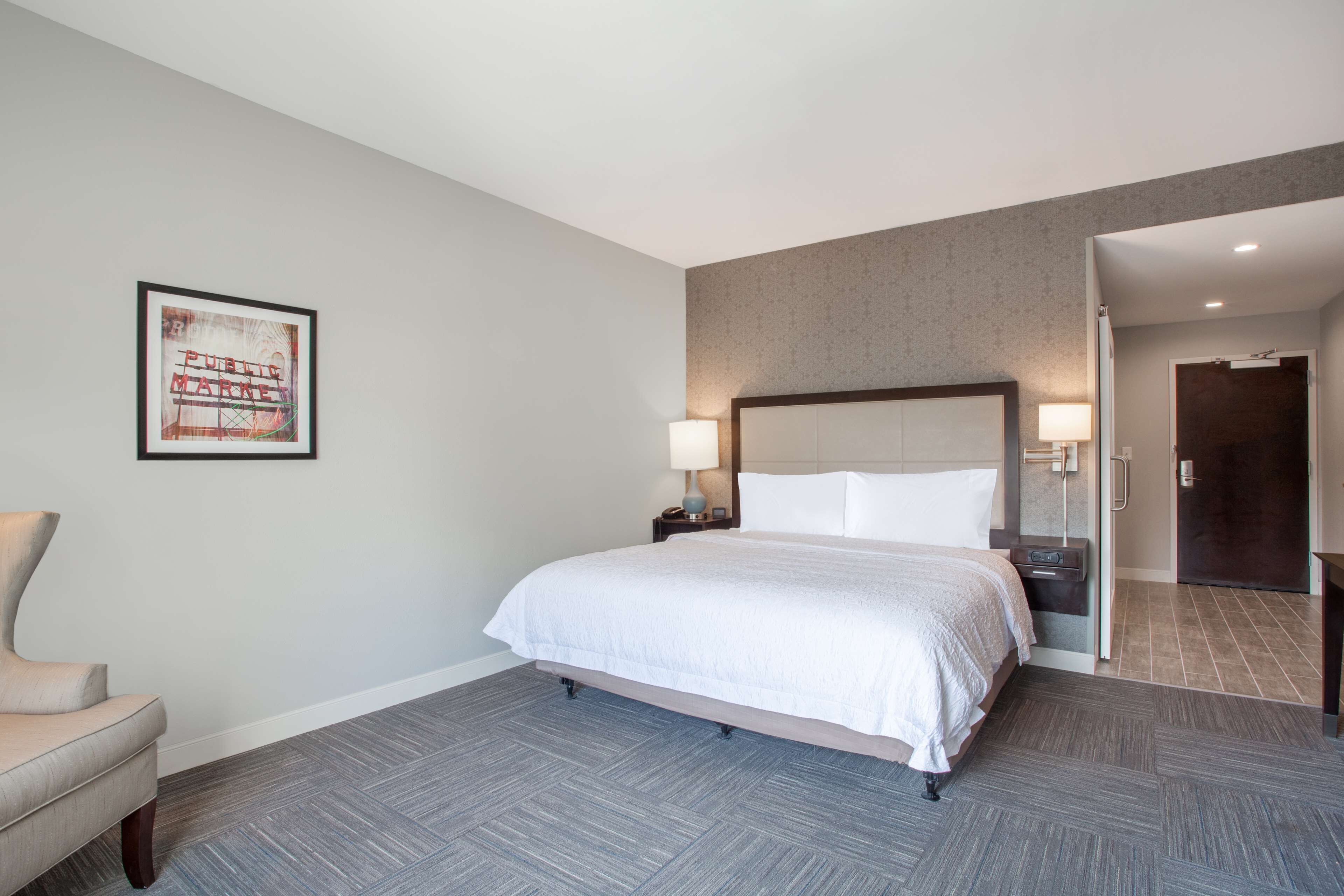 Une chambre d’hôtel avec un lit, une chaise, une table de chevet avec une lampe, un tableau encadré au mur et une porte donnant sur une autre pièce.