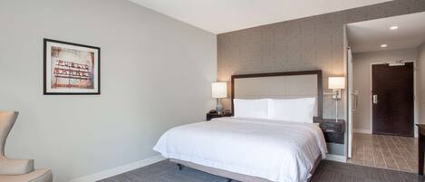 Une chambre d’hôtel avec un lit, une chaise, une table de chevet avec une lampe, un tableau encadré au mur et une porte donnant sur une autre pièce.