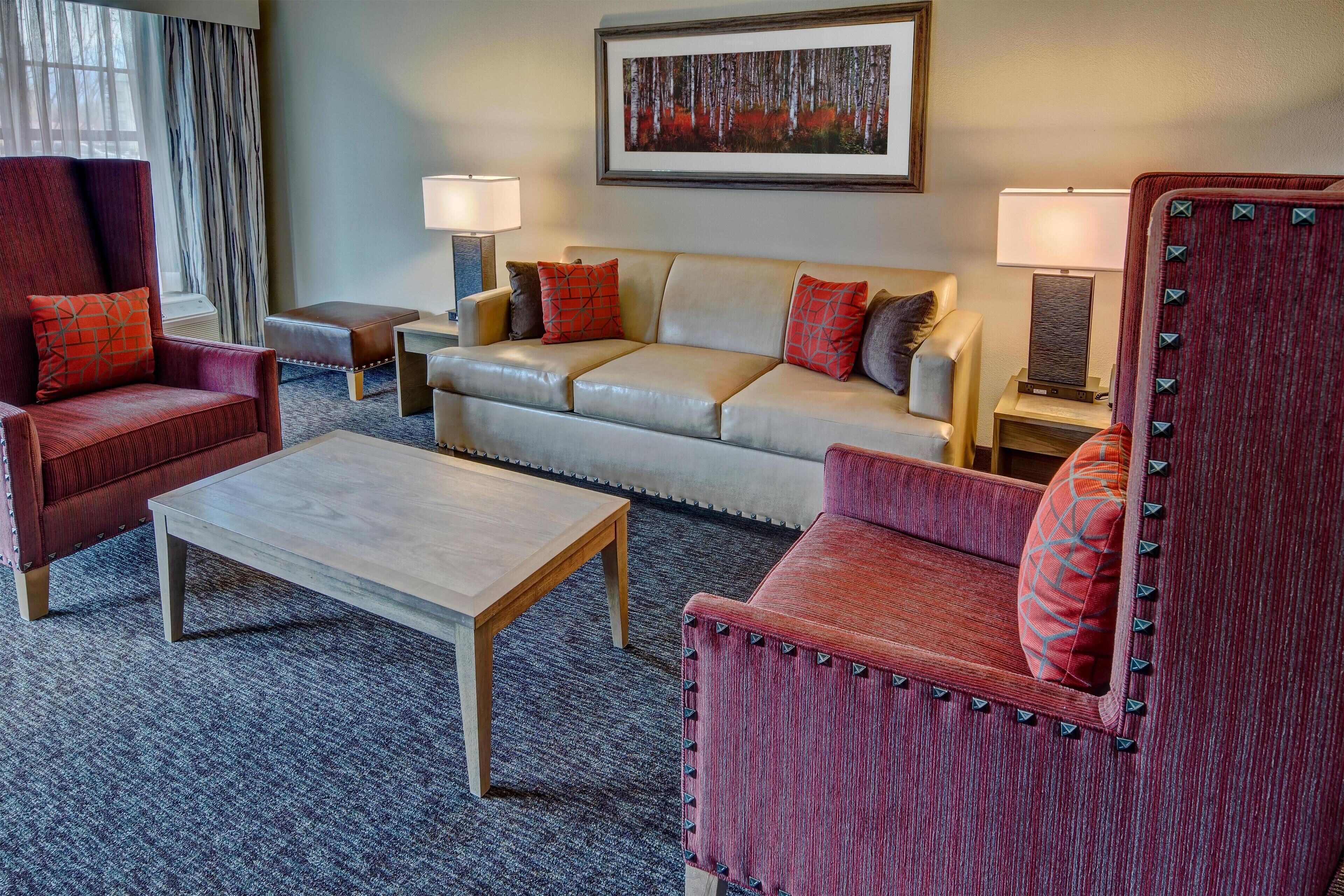 A hotel room with a red armchair, a beige sofa, a wooden coffee table, and two side tables with lamps.