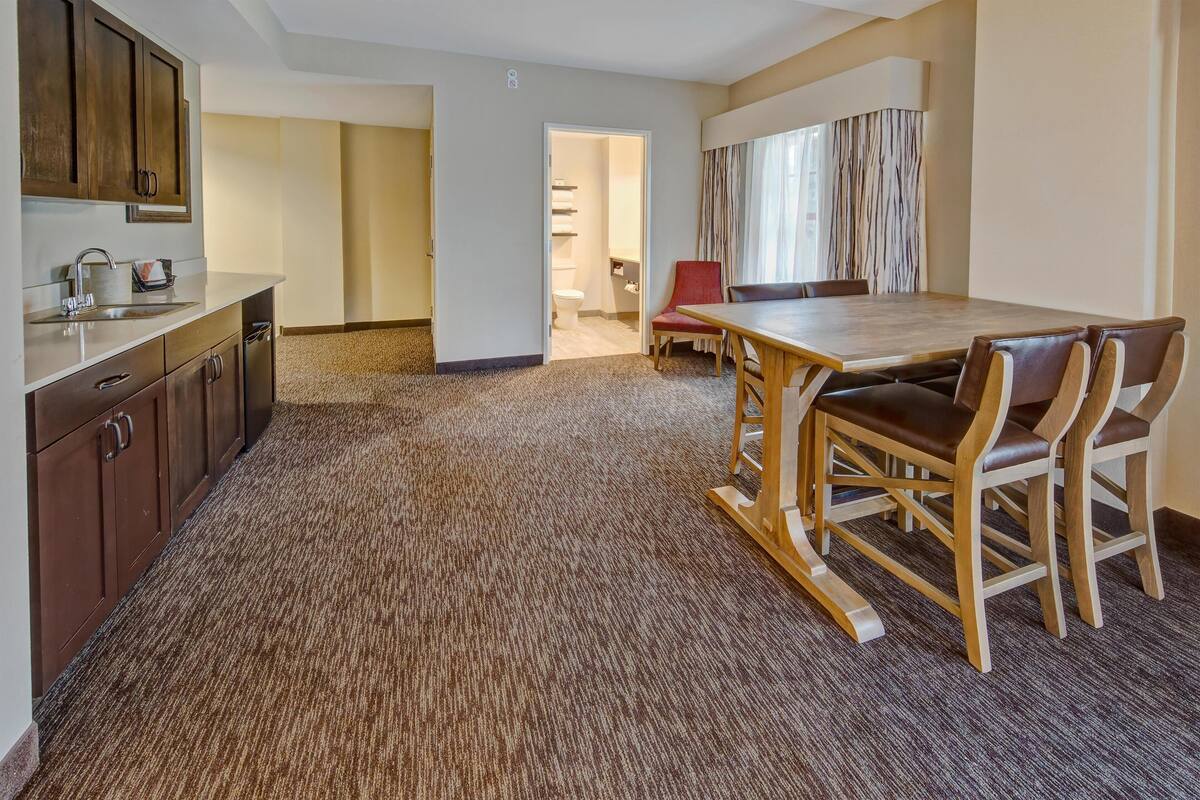 Habitación de hotel con una mesa de comedor de madera, cuatro sillas, una silla roja, una zona de cocina con armarios y un lavamanos, y suelo alfombrado.
