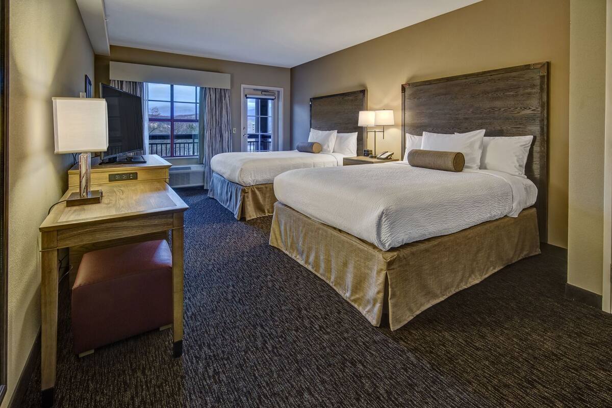 Habitación de hotel con dos camas, un escritorio, una silla, una lámpara y vistas al exterior.