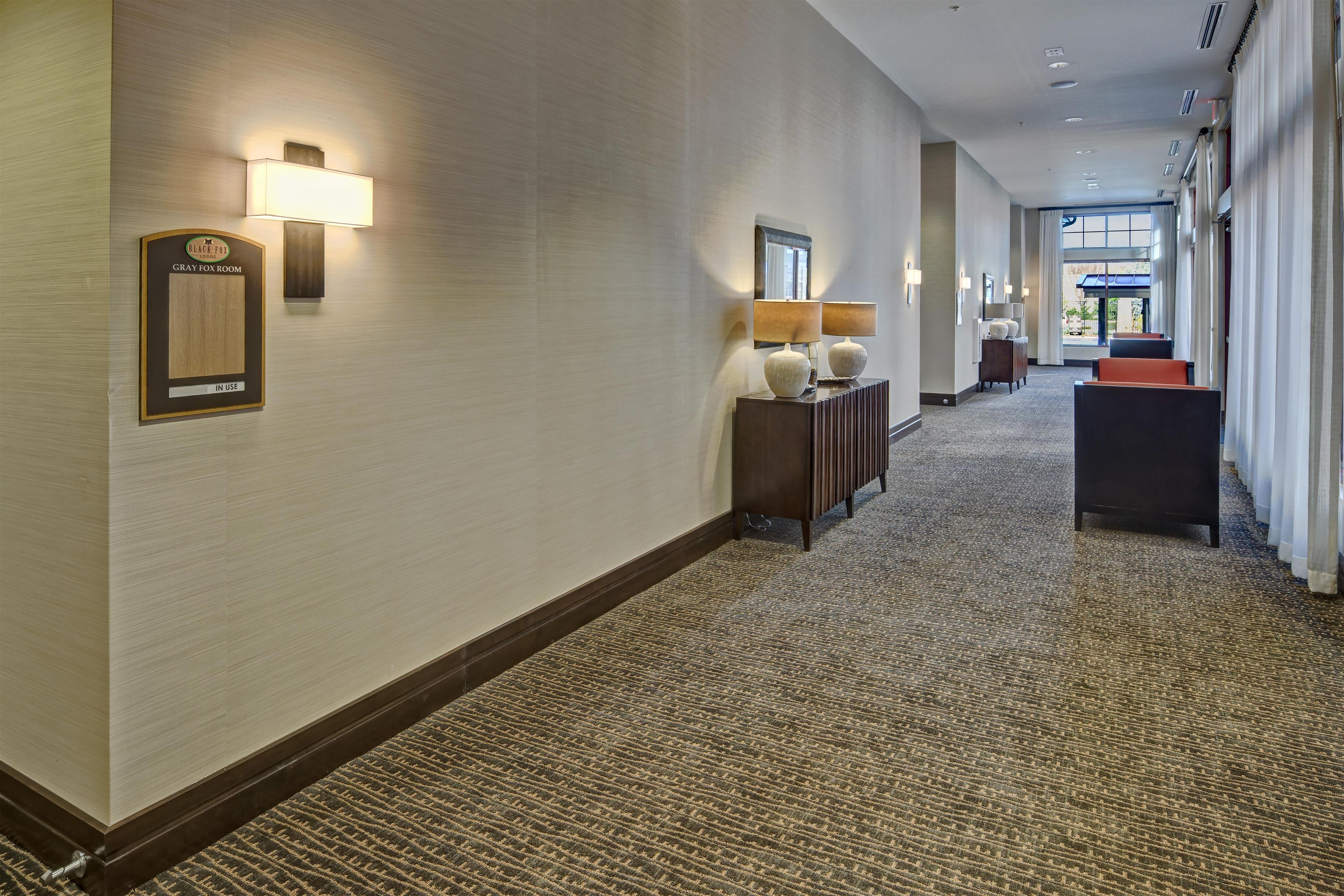 A hotel corridor with a carpeted floor, wall-mounted lamps, a sign indicating a room is in use, and a view of the outside through a window.