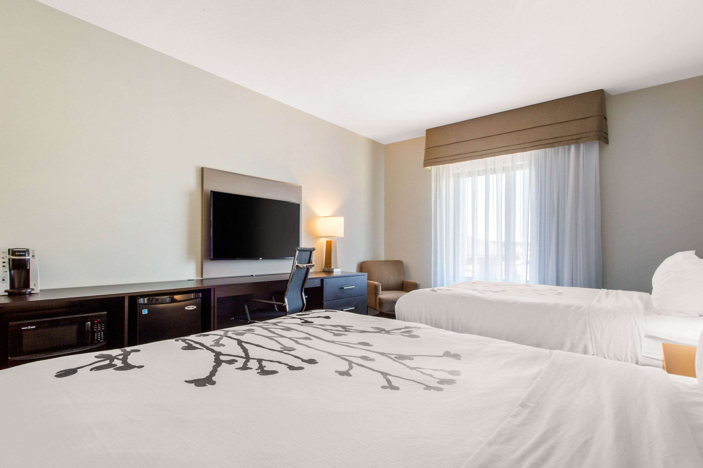 Ein Hotelzimmer mit einem großen Bett, einem Schreibtisch mit Flachbildfernseher, einem Sessel, einer Lampe und einem Fenster mit Vorhängen.