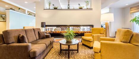 A modern lobby with a brown sofa, yellow armchairs, a patterned carpet, and a decorative plant centerpiece.
