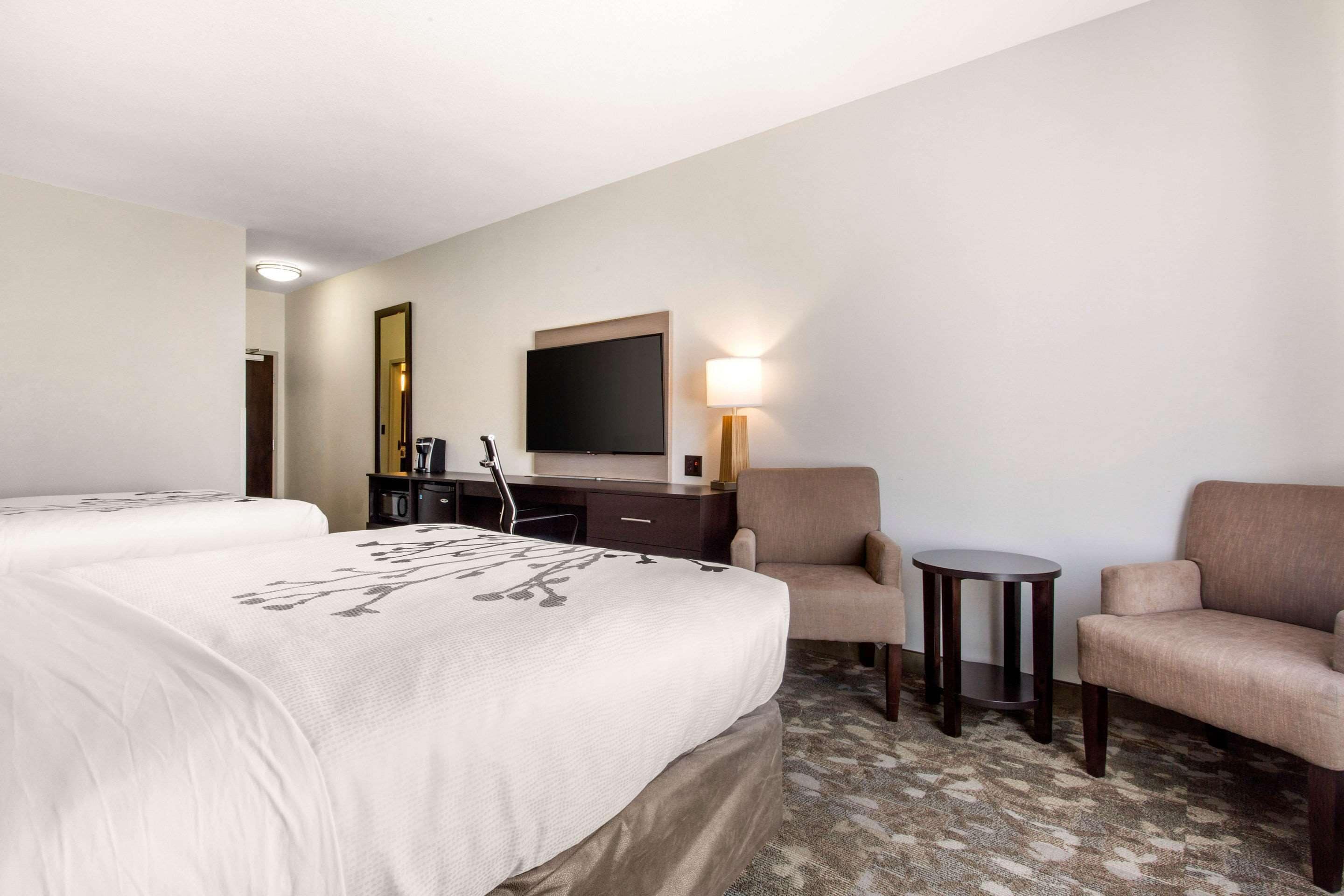A hotel room with two beds, a desk with a chair, a small table, a lamp, and a TV on the wall.