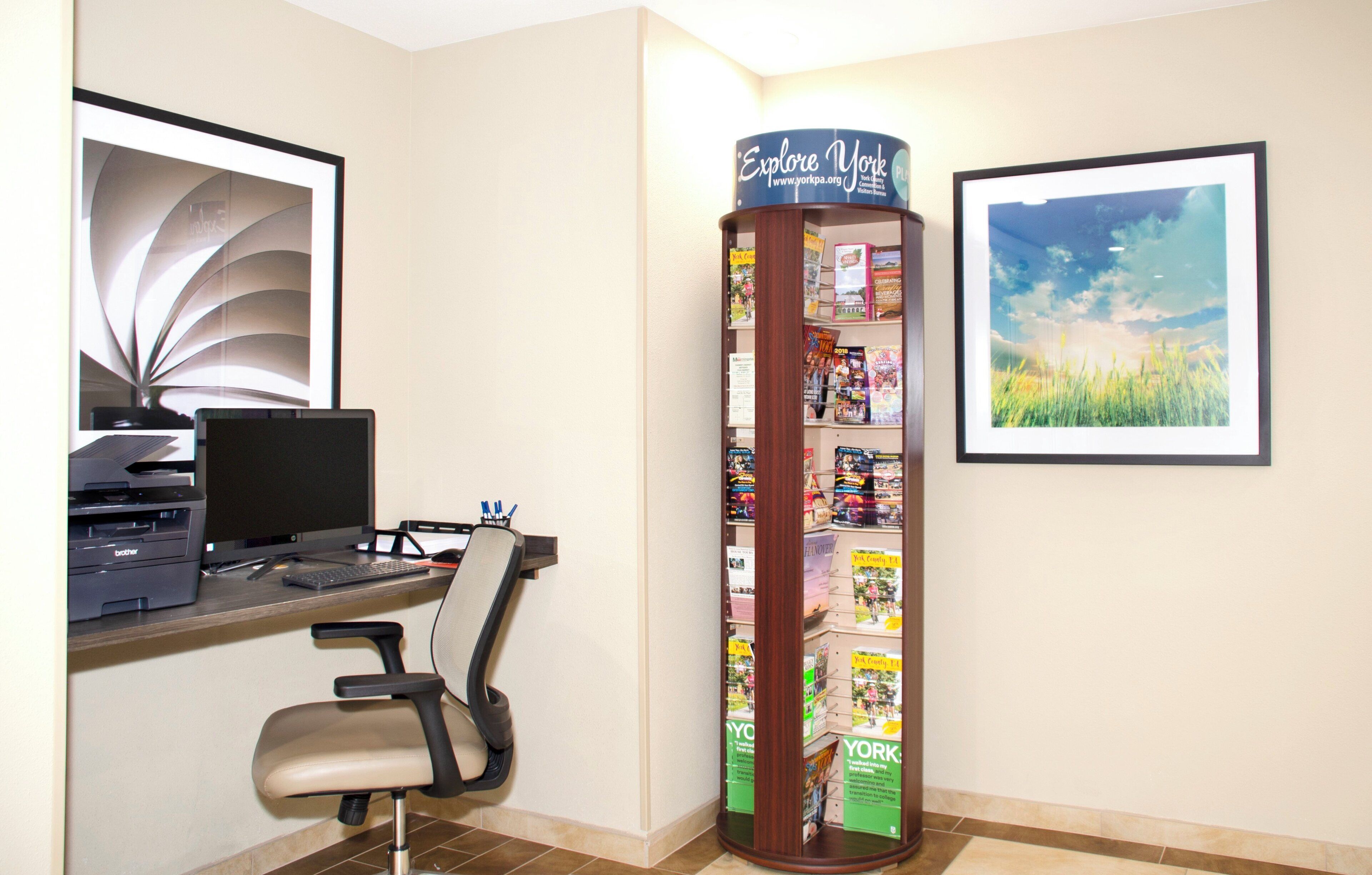 Un bureau avec un ordinateur, une chaise et une imprimante, un présentoir à magazines contenant des numéros du magazine « Explore York », et un tableau représentant un paysage, accroché au mur.