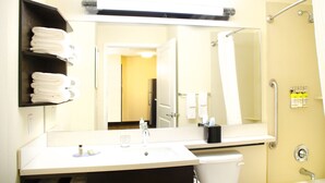 Een hotelbadkamer met een wastafel, spiegel en douche. Handdoeken liggen op een plank en toiletartikelen aan de muur.