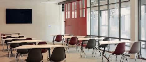 Ein Klassenzimmer mit Tischen, Stühlen, großen Fenstern und einem Flachbildfernseher an der Wand.