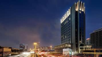 Un hotel moderno con un llamativo letrero iluminado, una calle concurrida con estelas de luz y una vista despejada de la fachada del edificio.