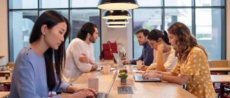 A group of people seated around a wooden table in a modern indoor setting with large windows and pendant lighting.