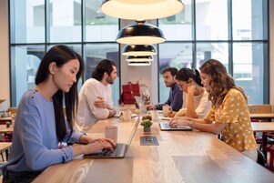 A group of people seated around a wooden table in a modern indoor setting with large windows and pendant lighting.