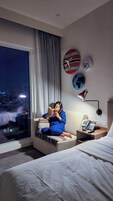 A hotel room with a large window, a bed, a bedside table with a lamp, a phone, and a wall with decorative plates.