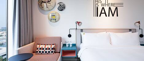 Ein Hotelzimmer mit einem Bett, einem Sessel, einem Schreibtisch und einer Wand mit Dekorationen, darunter eine Karte und ein Schild mit der Aufschrift 'THIS IS WHERE I AM'.