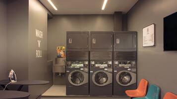 A laundry room with four washing machines, a small table with an iron, and two colorful chairs.