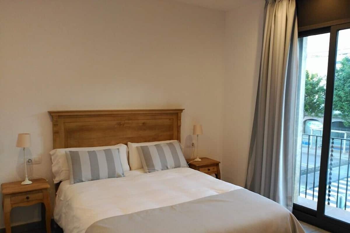 A hotel room with a wooden headboard, two bedside tables with lamps, a large bed with striped pillows, and a view of a balcony through a large window.