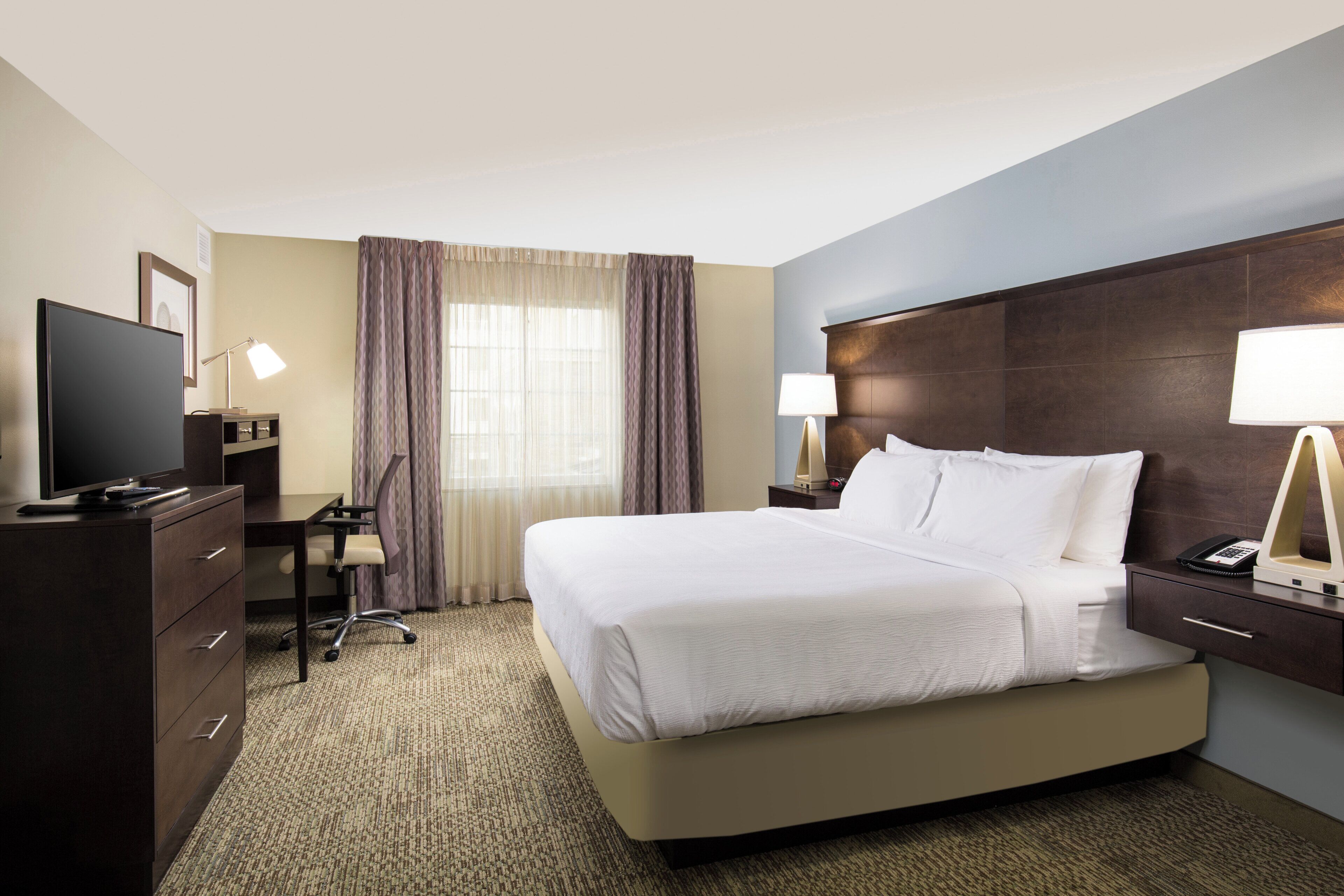 A hotel room with a bed, desk, chair, television, lamps, and a window with curtains.