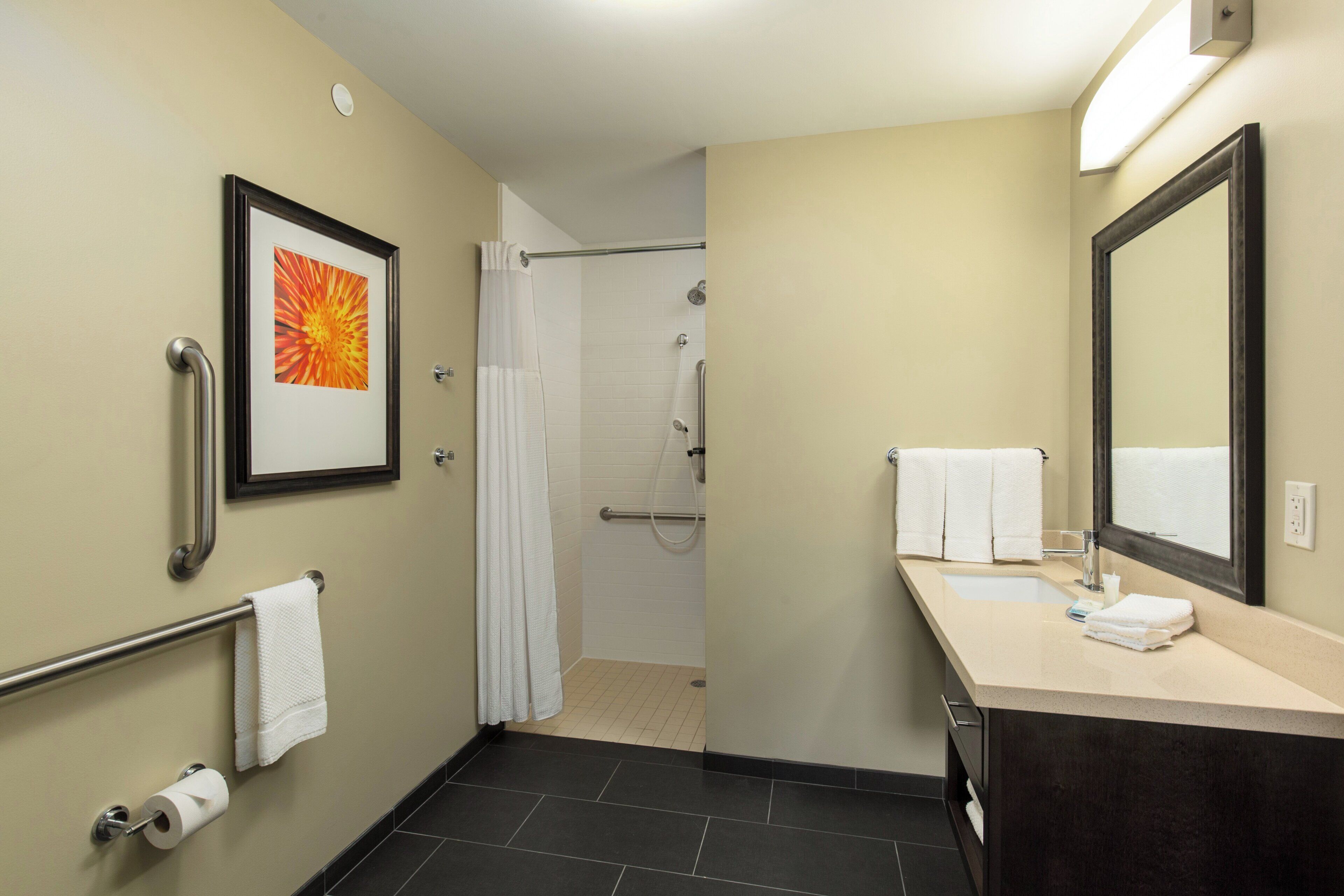 A hotel bathroom with a shower, toilet paper, a towel rack, and a sink with a mirror.