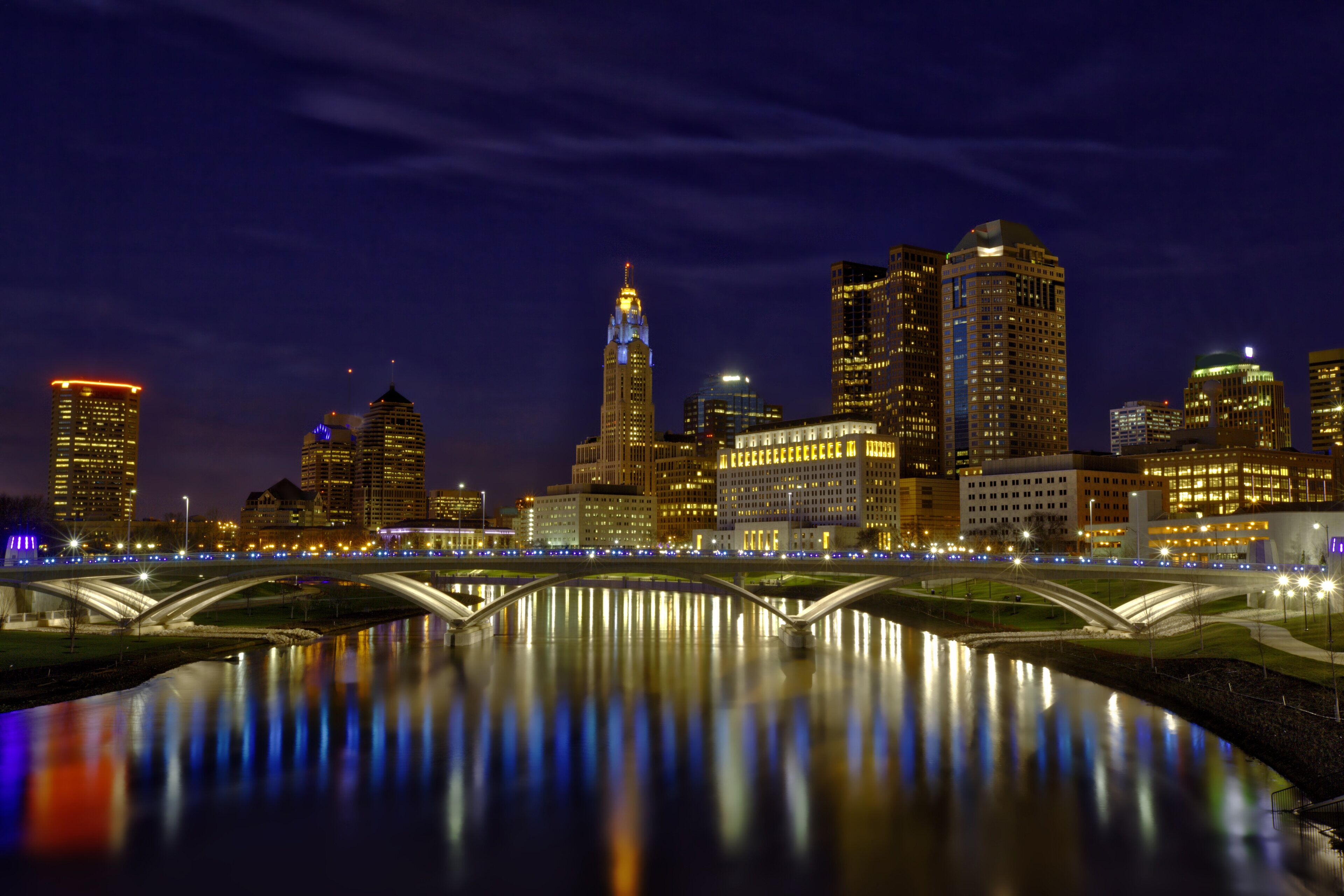 A nighttime cityscape with illuminated buildings and a bridge over a calm river.