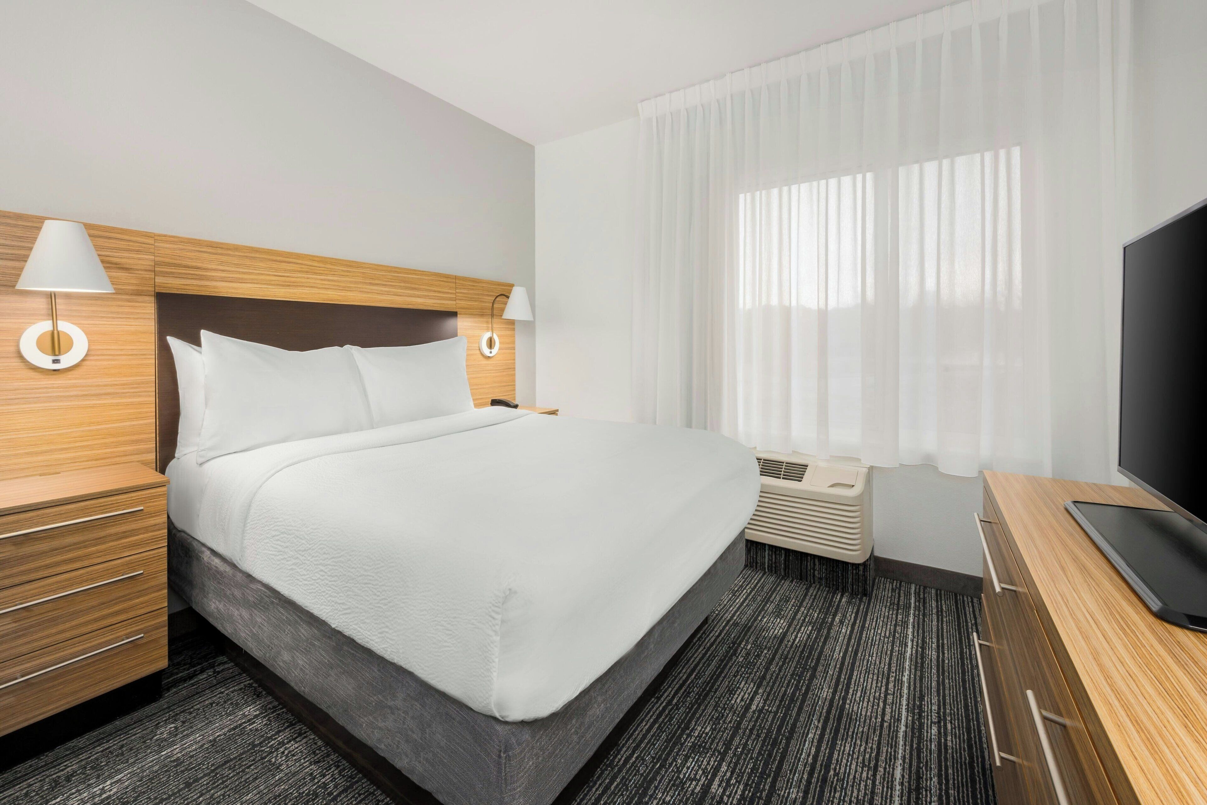 Una habitación de hotel con una cama grande, cabecera de madera, dos mesitas de noche con lámparas, un televisor y una ventana con cortinas de gasa.
