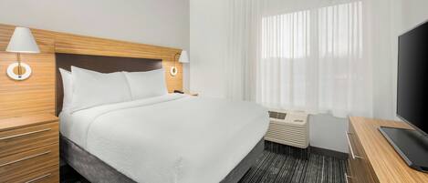A hotel room with a large bed, wooden headboard, two bedside tables with lamps, a TV, and a window with sheer curtains.