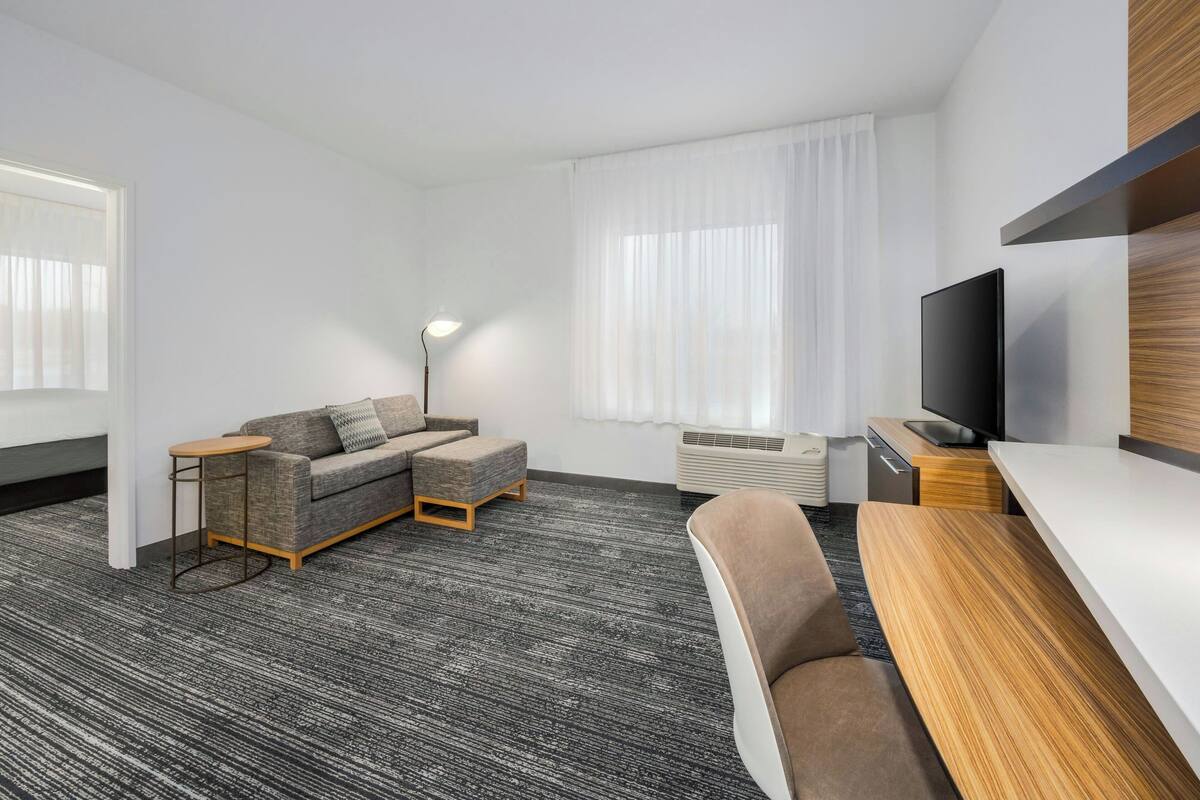 Quarto de hotel moderno com carpete cinza, sofá cinza, mesa lateral de madeira, televisão e uma grande janela com cortinas brancas.