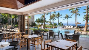 A restaurant with outdoor seating overlooking a pool and palm trees, featuring wooden tables and chairs.