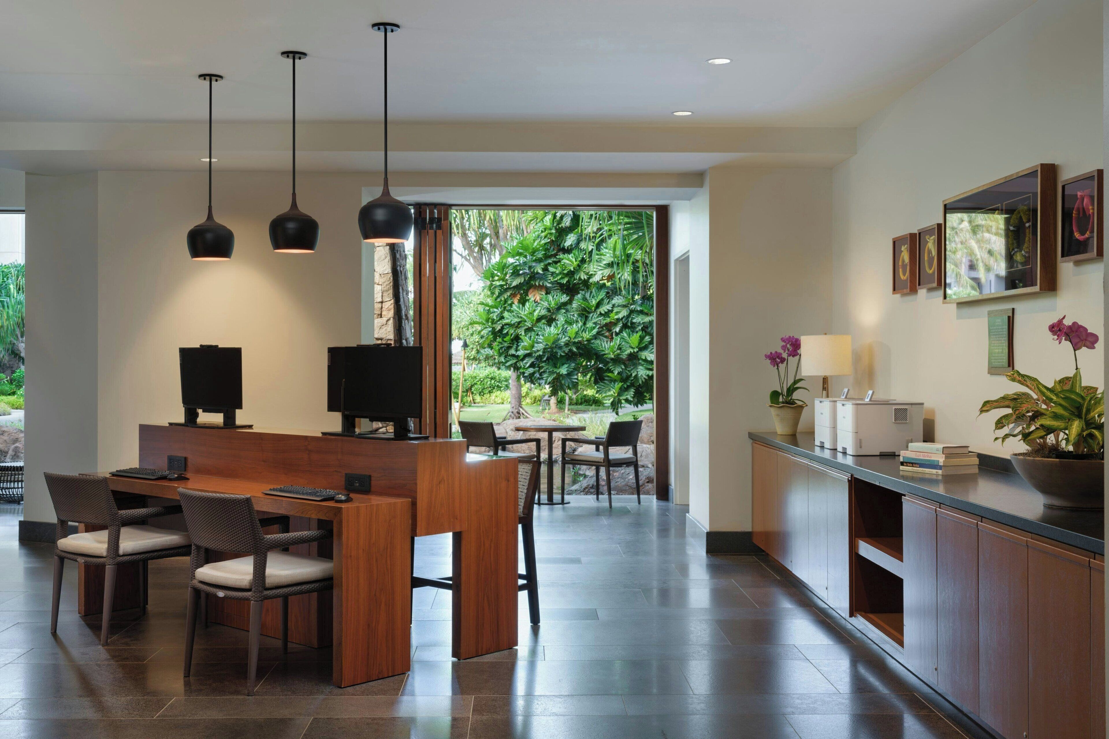A modern hotel lobby with a wooden reception desk, two black pendant lights, and a view of a lush garden through large windows.