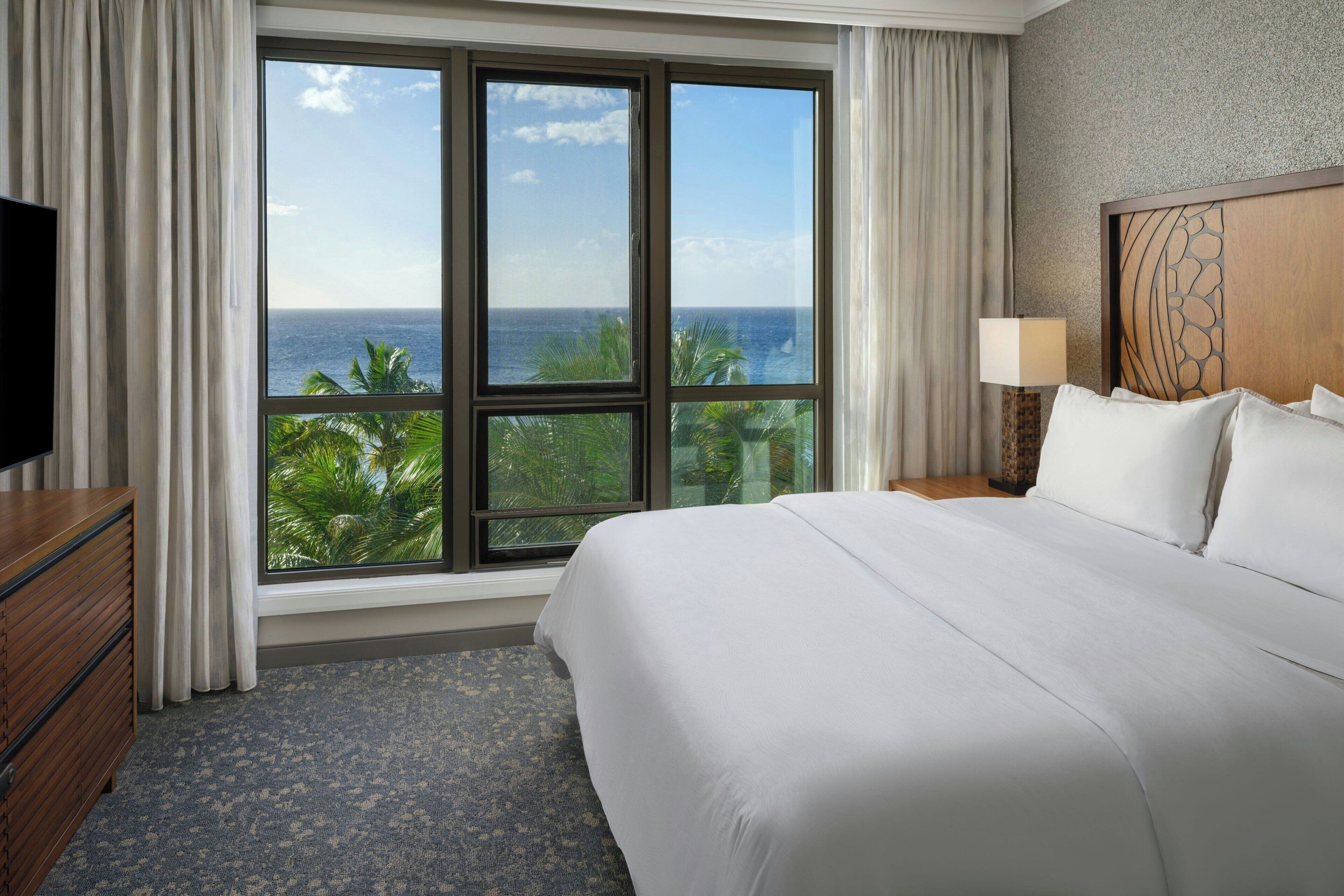 A hotel room with a large window offering a view of the ocean and palm trees.