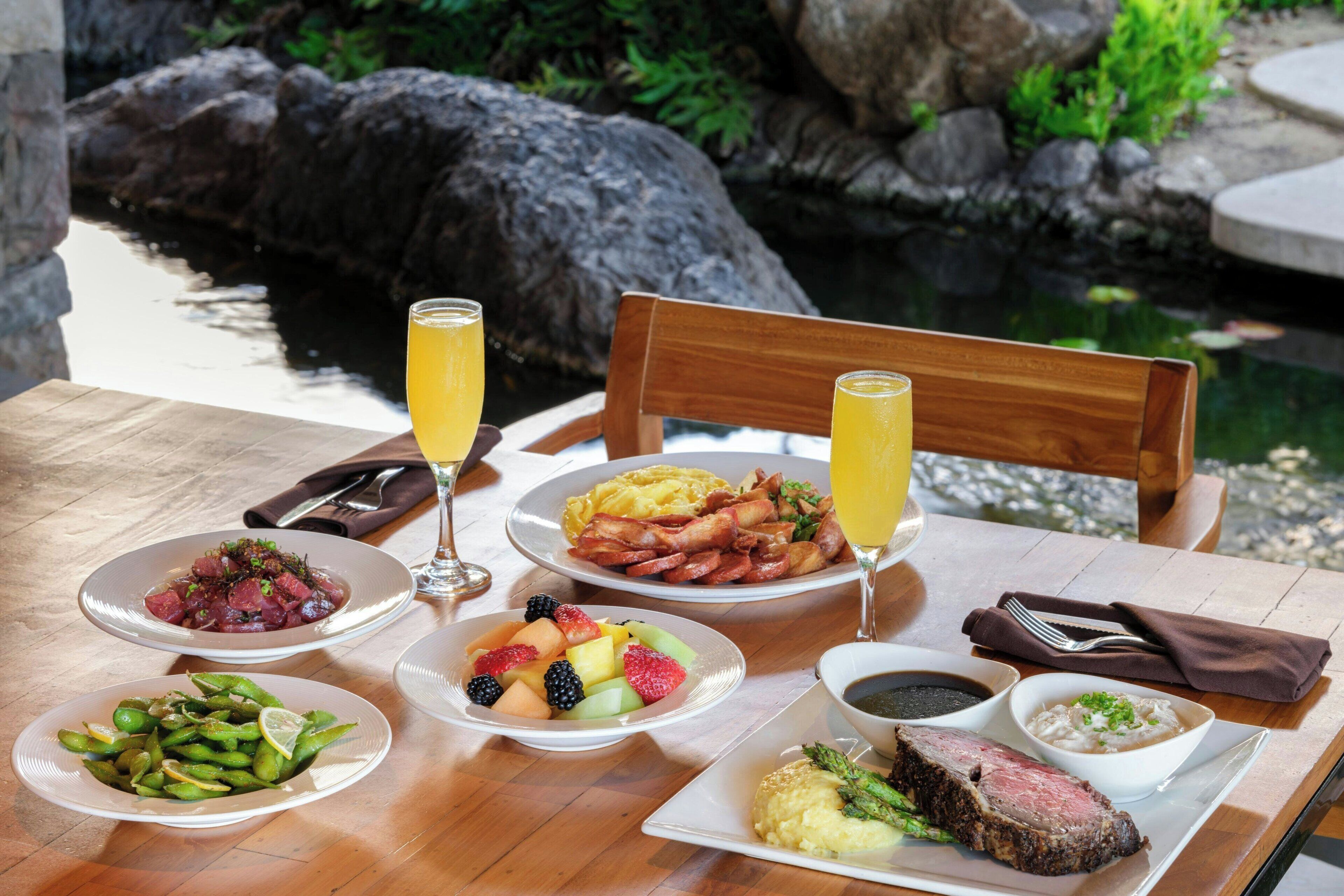 A table with various dishes including steak, fruits, and vegetables, set against a backdrop of a rocky garden and a pond.