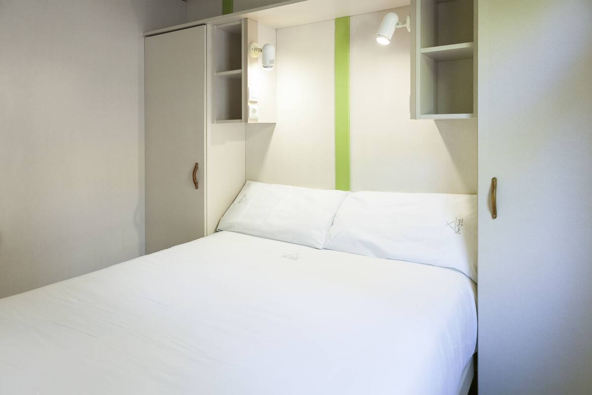 A neatly made bed in a room with a green stripe on the wall, a wardrobe, and a shelf with a light.