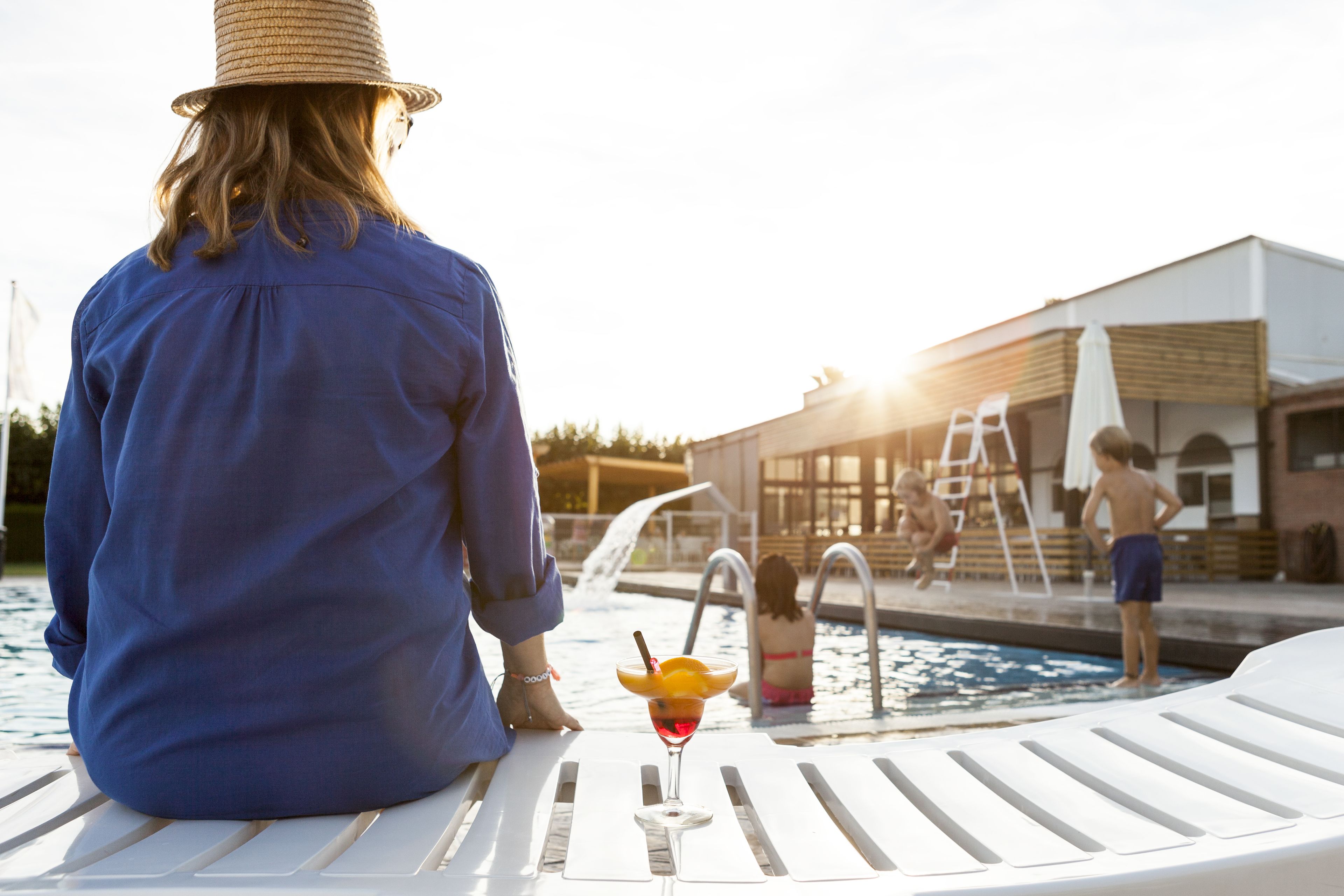 A person sitting by a poolside lounge chair with a drink in front, overlooking a swimming pool area with a building in the background.