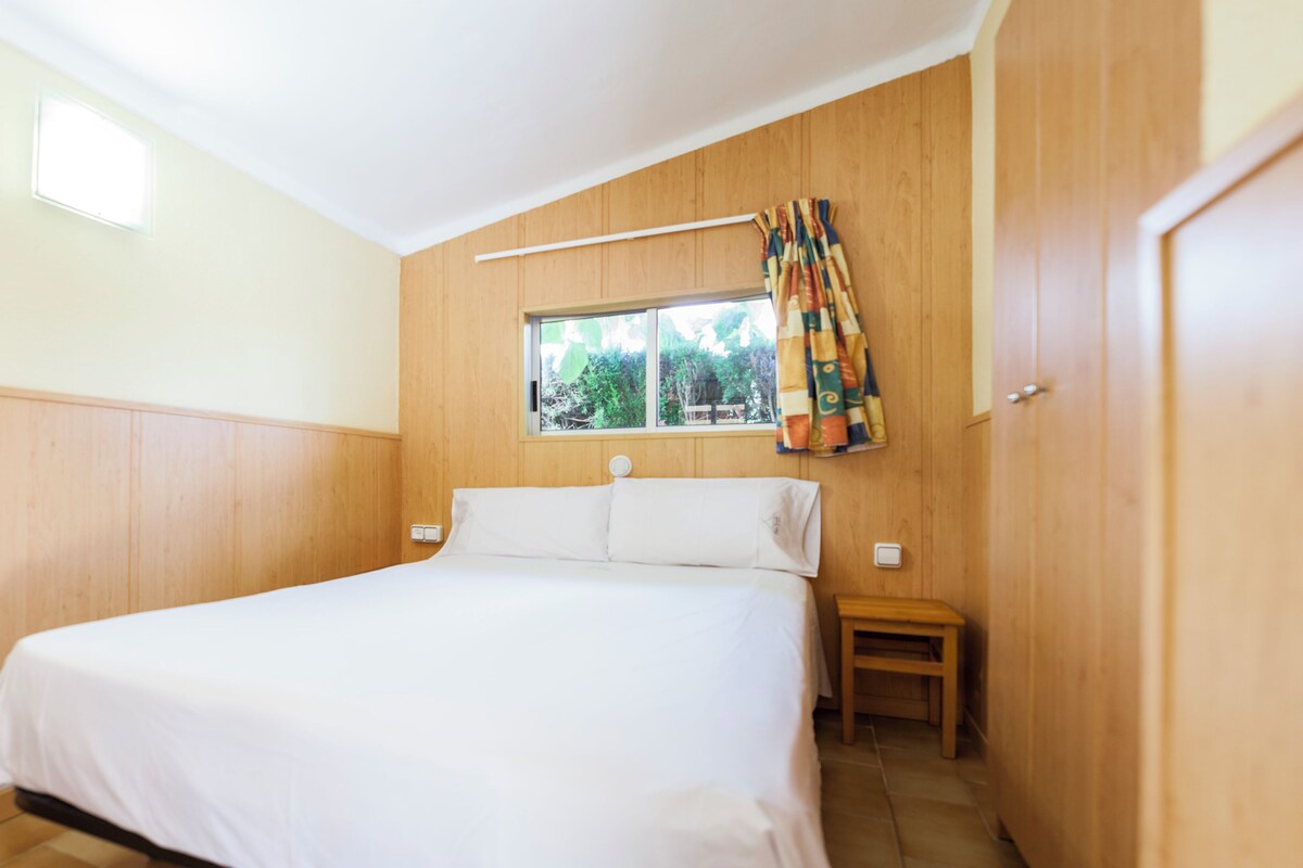 Un dormitorio con pared de paneles de madera, una ventana con vista a la vegetación, una cama con ropa blanca y una mesita de noche de madera.